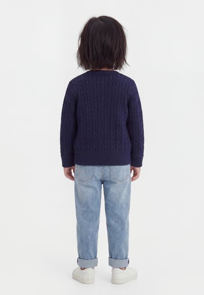 Dziecko stojące tyłem, ubrane w granatowy sweter w warkocze, jasnoniebieskie podwinięte jeansy i białe trampki na białym tle.