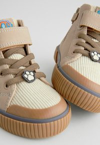 Scarpe alte beige chiare con tessuto cremoso testurizzato e lacci marroni. Presentano un accento a impronta di zampa e suole in gomma resistenti con scanalature.