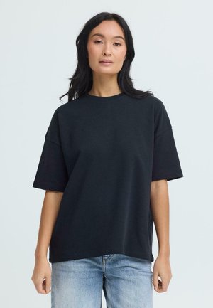 Femme aux longs cheveux foncés portant un t-shirt noir uni oversize et un jean bleu clair, debout devant un fond gris clair.