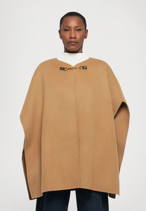 Camel-colored cape made of soft fabric, featuring a high collar, gold chain hardware, and wide, flowing design. Worn over a white top.