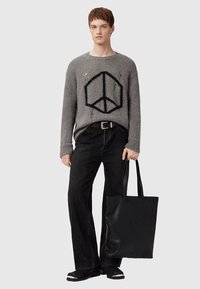 Nuori mies, jolla on yllään harmaa neulepaita, jossa on musta rauhansymboli, mustat housut ja kengät, pitää suurta mustaa kainalolaukkua sivullaan.