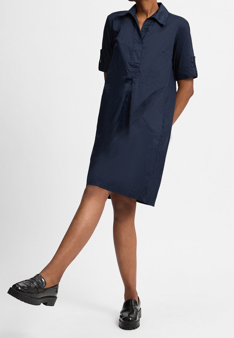 Frau in einem marineblauen knielangen Kleid mit kurzen Ärmeln und schwarzen, klobigen Loafers, die ein Bein leicht nach vorne hebt.