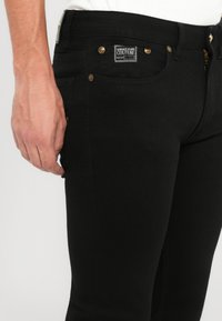 Melnas džinsos, kas izstrādāti ar šauru piegriezumu, izgatavoti no denima ar nelielu stiepjamo materiālu. Ievērojamas detaļas ir zelta detaļas un zīmola uzlīme uz jostasvietas.