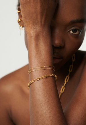 Persona de piel oscura con dos pulseras de oro en la muñeca, pendientes de aro dorados y un collar de cadena gruesa de oro, cubriéndose parcialmente la cara con el brazo.