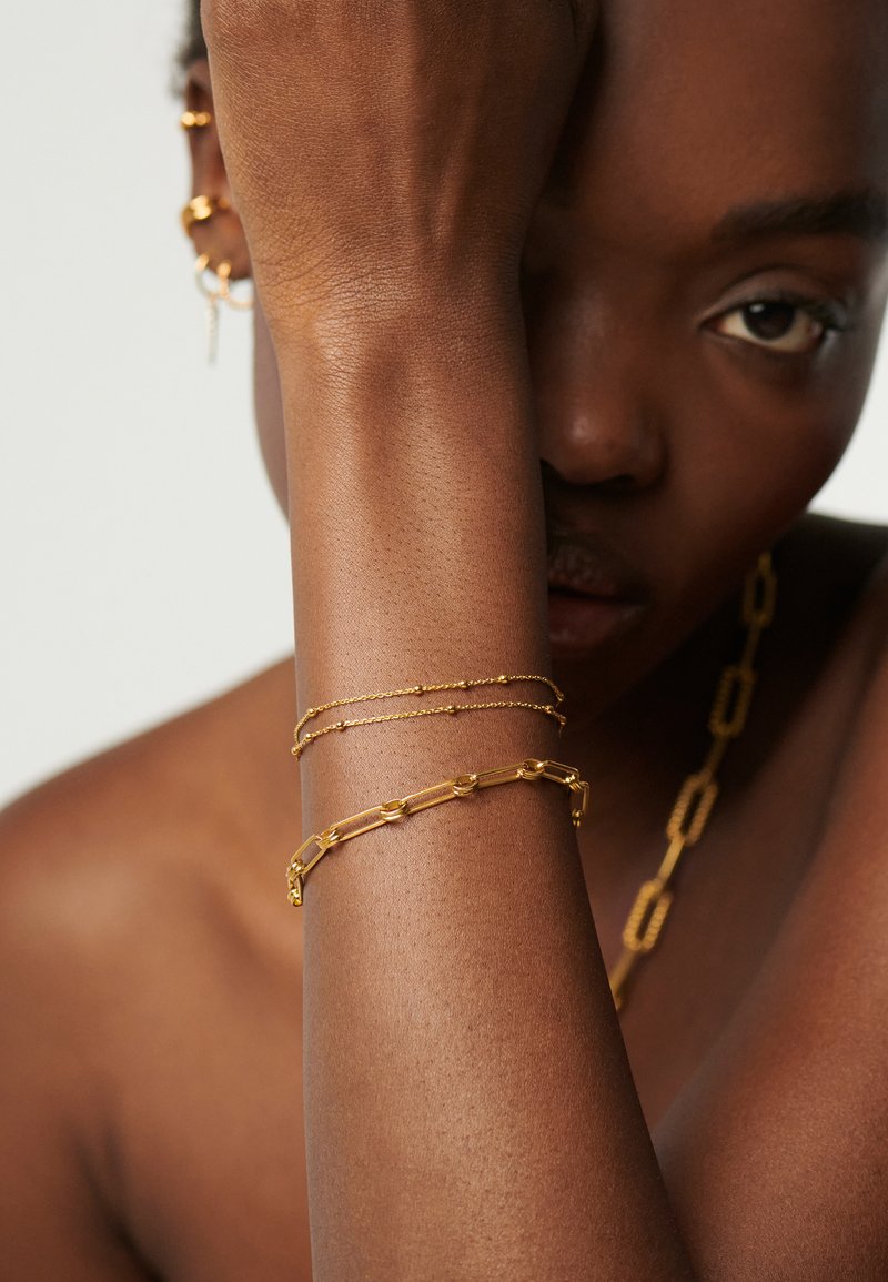 Osoba o ciemnej karnacji z dwoma złotymi bransoletkami na nadgarstku, złotymi kolczykami kołami i masywnym złotym łańcuchem na szyi, częściowo zasłaniająca twarz ramieniem.