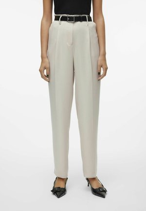 Pantalones beige de tiro alto con un corte entallado, que presentan un cinturón negro. Textura suave con pliegues frontales, combinados con zapatos negros de punta.
