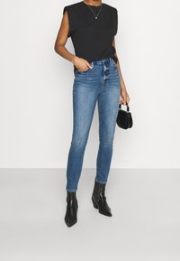 Svart ärmlös topp med axelvaddar, i kombination med tajta blå denimjeans och svarta klackade ankelskor. En liten svart handväska ingår.