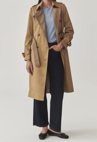 Béžový trenčkot s podšívkou v houndstooth vzoru, dvouřadové knoflíky a opasek v pase, nošený přes světle modrý top a tmavé džíny.