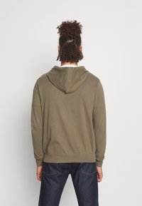 Sudadera con capucha de color verde oliva, de corte holgado, mangas largas y ribete acanalado. Detalles de costuras visibles en la parte trasera. Combinada con jeans oscuros.