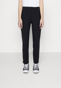 Pantalon noir fuselé en tissu extensible avec une texture lisse, doté d'une taille mi-haute et de passants de ceinture standards. Assorti à des baskets noires.