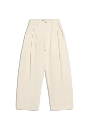 Junger Mann mit roter Kappe, weißem Langarmhemd und weiten cremefarbenen Hosen steht zwischen Edelstahl-Kaffeemaschinen in einem Café.