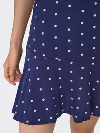 Abito da sera blu navy con pois bianchi, orlo svasato, tessuto leggero e maniche corte. Primo piano di una mano che afferra la gonna.