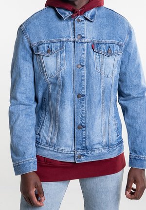 Uomo con giacca di jeans azzurro chiaro sopra una felpa con cappuccio rossa, abbinata a jeans azzurri, in piedi contro uno sfondo bianco.