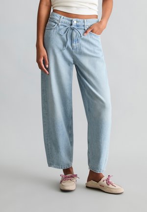 Frau trägt hellblaue Jeans mit weitem Bein und Kordelzug in der Taille, ein weißes bauchfreies Top und beige Mokassins mit rosa Schnürsenkeln.