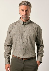 Hombre de mediana edad con cabello y barba canosos, vistiendo una camisa de botones de manga larga con estampado metida dentro de pantalones oliva, de pie frente a un fondo liso.
