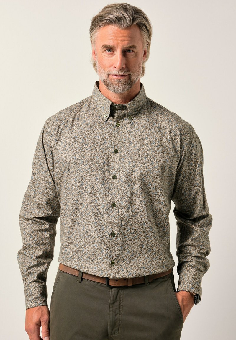 Hombre de mediana edad con cabello y barba canosos, vistiendo una camisa de botones de manga larga con estampado metida dentro de pantalones oliva, de pie frente a un fondo liso.