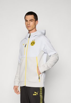 Lys grå zip-up hoodie med en gul lynlås og logo. Har hætte, sidelommer og kontrasterende mørkegrå bukser med en gul detalje.