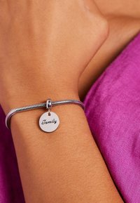 Zilveren armband met een gestructureerde band, met een ronde hanger gegraveerd met "Familie." De achtergrond is paarse stof.