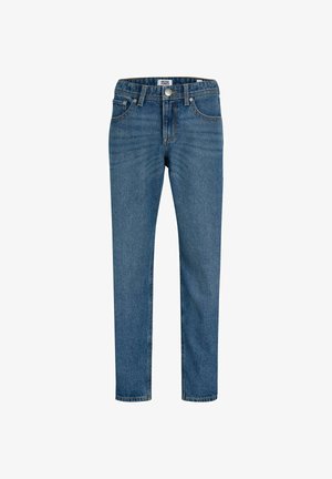 Garçon souriant aux cheveux bouclés portant une chemise à carreaux noire et grise, un jean bleu ample et des bottes lacées beiges, debout les mains dans les poches.