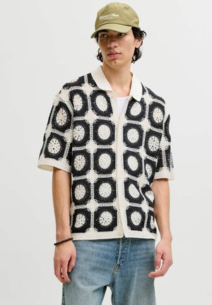 Junger Mann, der ein schwarz-weiß gehäkeltes Kurzarmhemd, hellblaue Jeans, eine olivgrüne Mütze und ein schwarzes Armband am linken Handgelenk trägt.