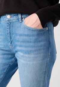 Šviesiai mėlynos džinsinės kelnės su slim fit fasonu, turinčios priekinę kišenę ir sidabrinius aksesuarus diržo srityje. Lygi tekstūra, kasdienis dizainas.