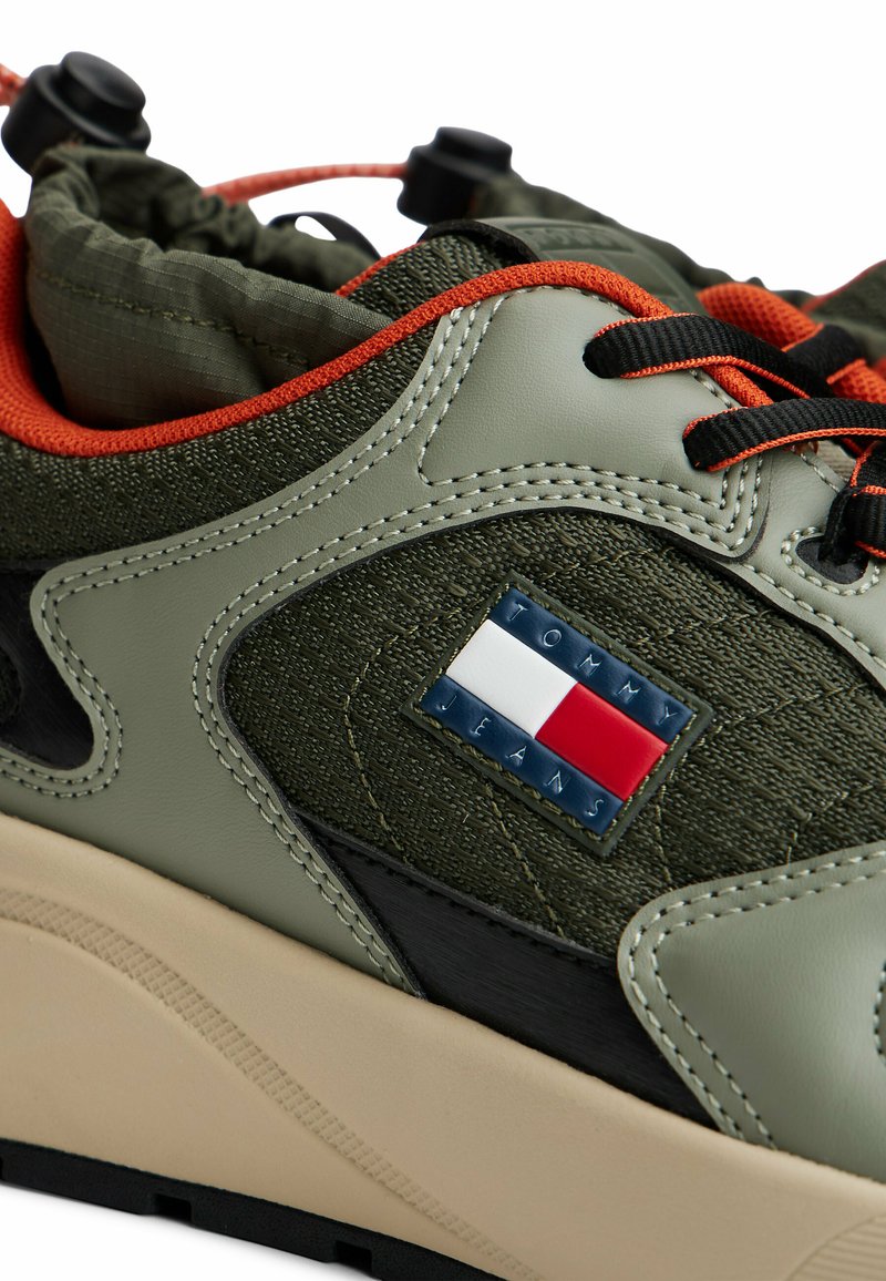 Green and black sneaker with plush fabric, textured overlays, orange accents, and a Tommy Jeans logo patch. Features a thick beige sole.