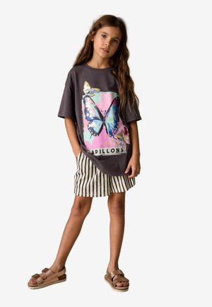 Graues übergroßes T-Shirt mit Schmetterlingsmotiv in Pink und Blau, kombiniert mit gestreiften Shorts. Braune Sandalen mit Riemen und leichtem Plateau.