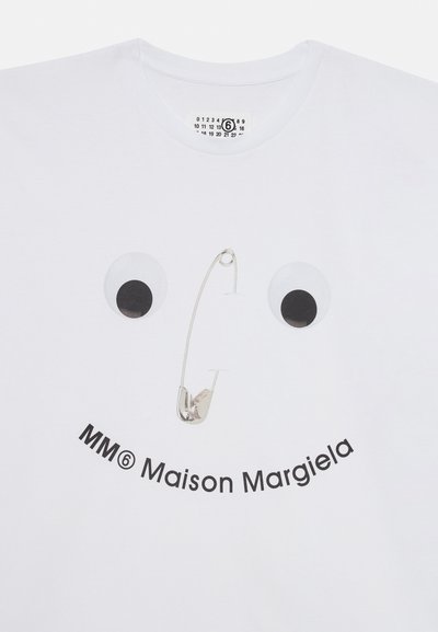Balta kokvilnas T-krekls ar divām lielām melnām acīm un sudraba drošības adatu, ar uzrakstu "MM6 Maison Margiela" izdrukātu izliekta stilā.