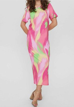 Femme portant une robe midi à motifs abstraits rose, verte et jaune avec des manches courtes et des sandales à talons bloc beiges, se tenant avec les mains derrière le dos.