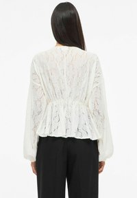 Biała koronkowa bluzka z baskinką, długimi bufiastymi rękawami i marszczoną talią. Tkanina ozdobiona teksturowanym, kwiatowym wzorem.