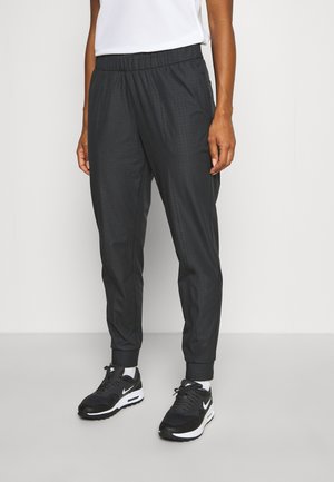Person trägt schwarze Jogginghose mit elastischem Bund, weiße Socken und schwarze Nike-Sneaker, steht vor einem schlichten, hellen Hintergrund.