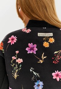 Czarny sweatshirt w kwiatowy wzór z jaskrawymi różowymi, fioletowymi i żółtymi kwiatami. Żebrowany kołnierz z metką oznaczającą "EFEKT MOTYLA". Miękka tekstura.