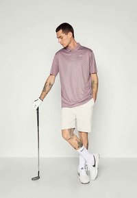 Μωβ κοντομάνικο πουκάμισο πόλο με κυκλικό μοτίβο, σε συνδυασμό με λευκά σορτς. Μια γαλλική ρακέτα κρατιέται στο δεξί χέρι, με λευκές κάλτσες και παπούτσια.