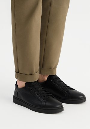 Baskets en cuir noir avec une finition texturée, design à lacets et semelle en caoutchouc noir plate, associées à un pantalon olive vert à revers.
