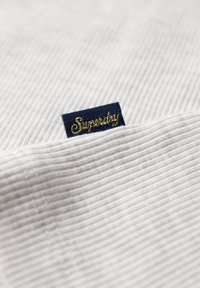 Tejido de rayas gris con una etiqueta azul marino que presenta "Superdry" en texto cursivo dorado, parcialmente doblado, revelando la textura y los detalles de diseño.