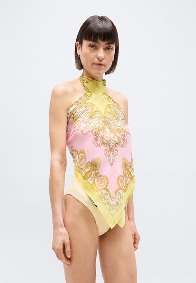 Γυναίκα με κοντά σκούρα μαλλιά, που φοράει ένα  κίτρινο και ροζ σχέδιο τοπ με μαντήλι τύπου halter και μπεζ σλιπ με ψηλή κοπή, στέκεται μπροστά σε απλό λευκό φόντο.