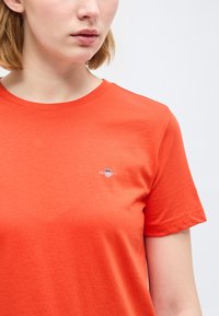 Γυναίκα που φοράει απλό πορτοκαλί κοντομάνικο μπλουζάκι με μικρό λευκό λογότυπο ασπίδας στο αριστερό στήθος, μπεζ φόντο.