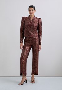 Traje metálico burdeos con chaqueta ajustada y corta y pantalones de pierna recta. La tela presenta un patrón sutil y una textura brillante. Tacones abiertos negros.