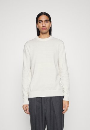 Maglione a maniche lunghe di colore crema chiaro, in una morbida trama a maglia, con scollo rotondo, abbinato a pantaloni scuri e larghi.