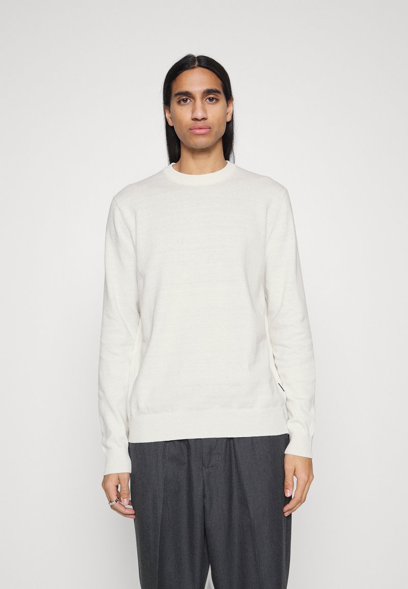 Maglione a maniche lunghe di colore crema chiaro, in una morbida trama a maglia, con scollo rotondo, abbinato a pantaloni scuri e larghi.