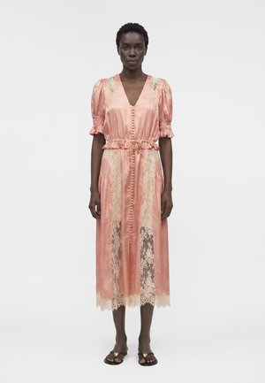 Kobieta stoi, ubrana w różową satynową sukienkę midi z koronkowymi panelami, bufkami, guzikami z przodu oraz czarne sandały ze złotymi ozdobami w kształcie serc.