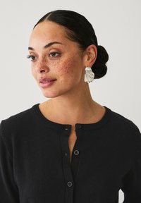 Orecchini a statement in tono argento con un design ondulato e texturizzato. Il modello indossa un cardigan nero con chiusura a bottoni e scollo rotondo.