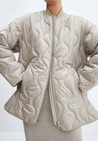 Beige quiltet bomberjakke med bølgende tekstur, ribbet krave og sølvfarvet lynlås, der har en afslappet pasform og brede ærmer.