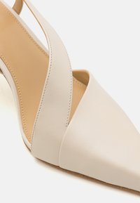 Pointed-toe pump in cream leather with an angular cutout design. Features a slender strap and a beige outsole with subtle texture.
