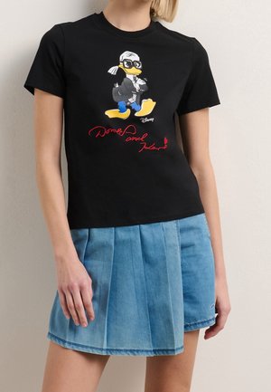 Femme portant un t-shirt noir avec un dessin de Donald Duck en lunettes de soleil et une jupe en jean plissée bleue.