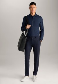Marineblauwe longsleeve shirt, slim-fit donkere broek, witte sneakers en een zwarte leren tote bag. Gladde textuur met knoopdetails.