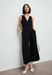 Zwarte mouwloze jumpsuit met een diepe V-hals, een ceintuur om de taille, een brede broekspijp en zijzakken. Een paar lichtgekleurde open teen-hakken.