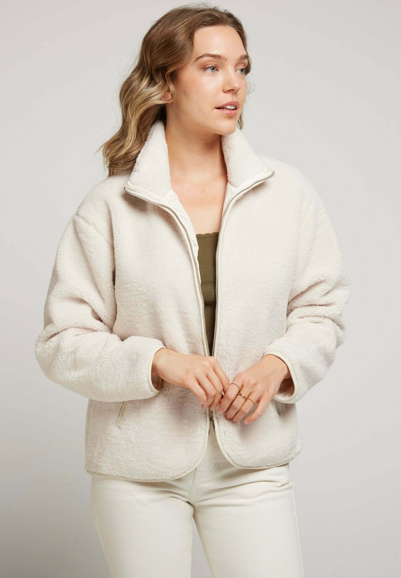 Veste en polaire beige clair avec un col haut, une fermeture éclair à l'avant et des poches latérales ; texture douce avec une coupe décontractée, assortie à un haut sombre.