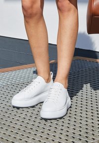 Jambes portant des baskets blanches épaisses à lacets, debout sur une grille métallique, partie d'un sac marron visible sur le côté droit.