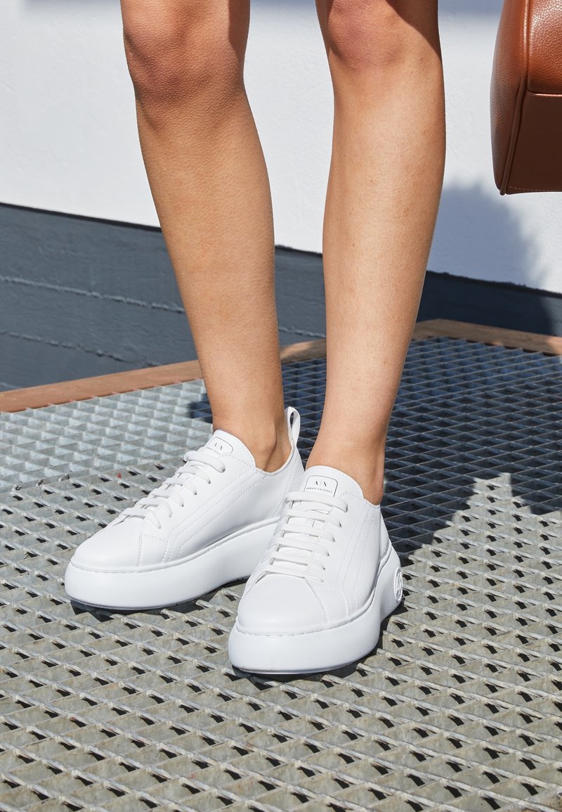 Jambes portant des baskets blanches épaisses à lacets, debout sur une grille métallique, partie d'un sac marron visible sur le côté droit.